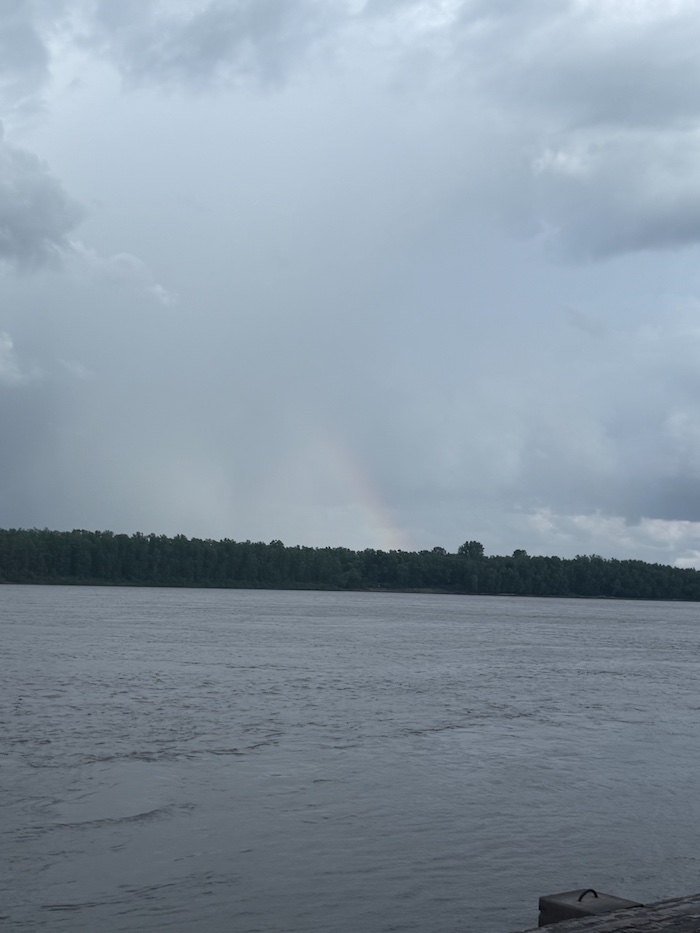 Picture of the river with a rainbow above the tree line in the distance. This is a great portrayal of savoring life and slow living.